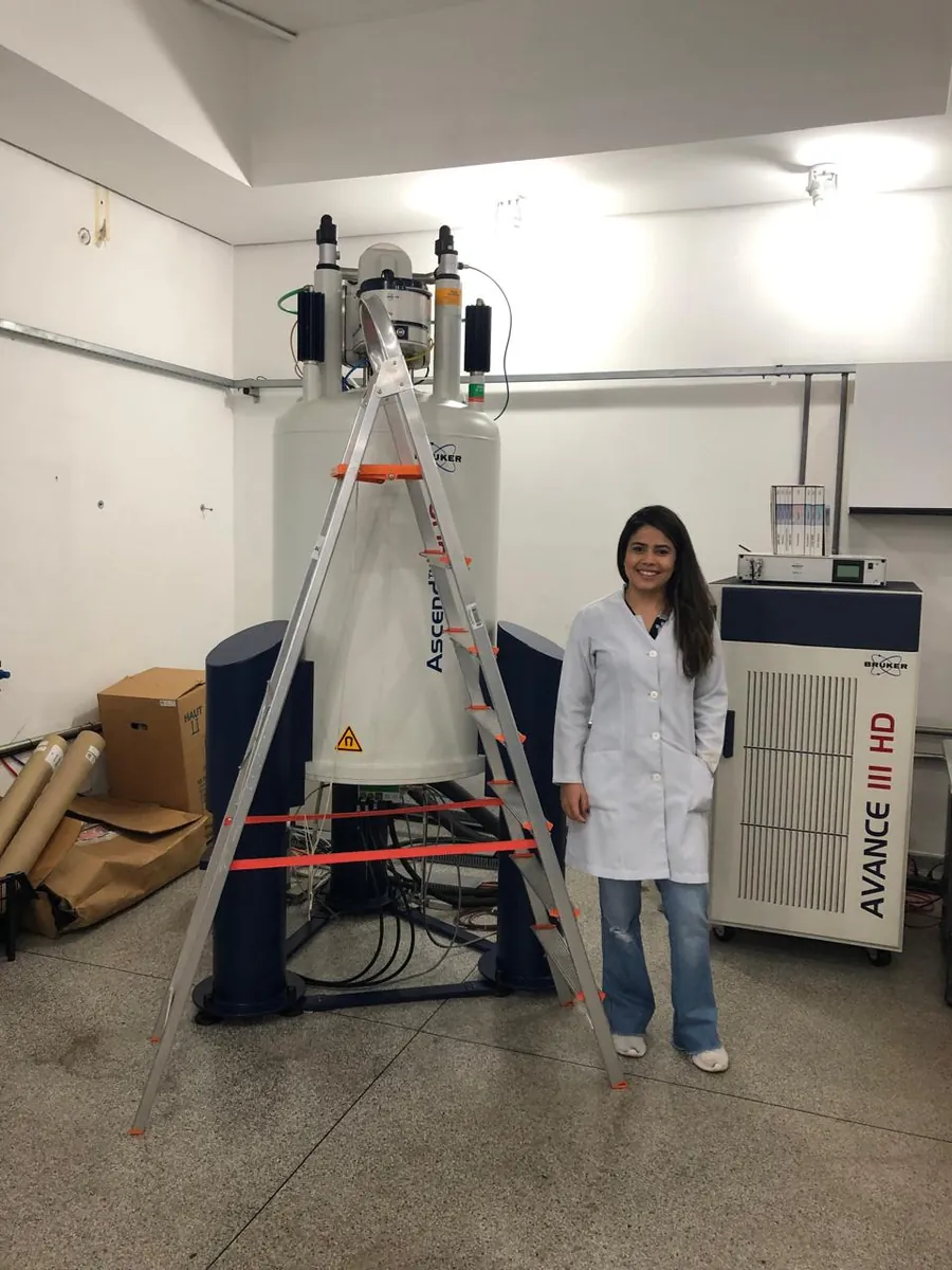 Williara Queiroz ao lado de um espectrômetro de ressonância magnética nuclear Bruker Avance III HD